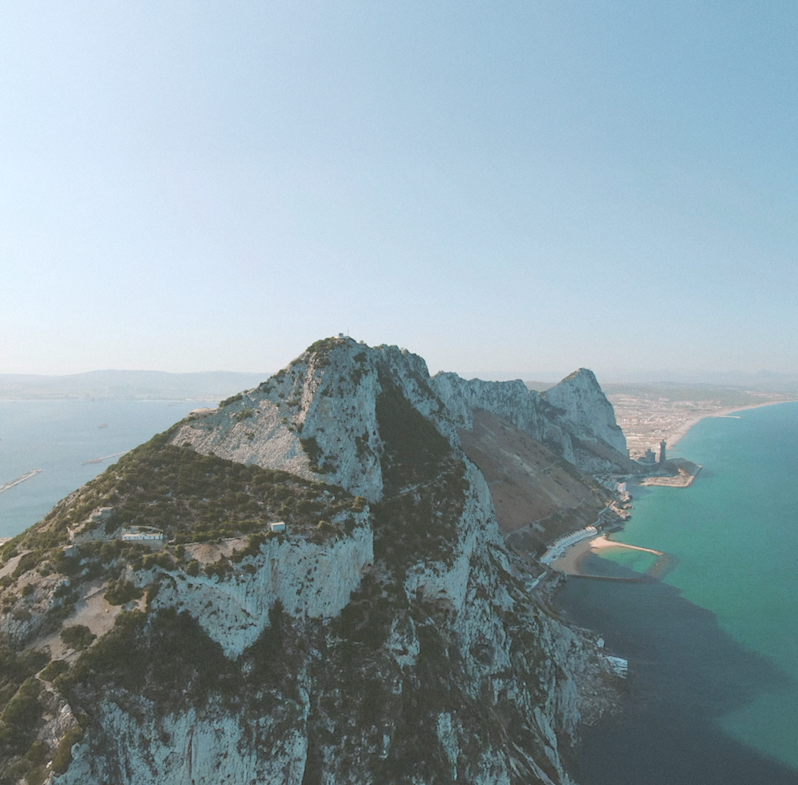 An aerial drone view looking down the rugged ridge of the Rock of Gibraltar surrounded by blue ocean water.