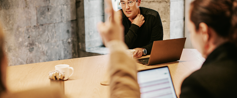 Colleagues collaborating during a business meeting in an industrial-style boardroom, showing a team member gesturing while others listen and work on their laptops.