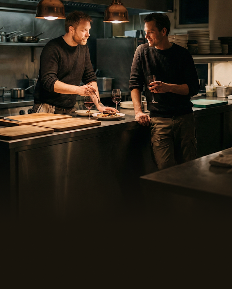 Professional chefs tasting a new gourmet menu item in a commercial kitchen setup during an evening service, holding wine glasses and showcasing a behind-the-scenes premium culinary experience.