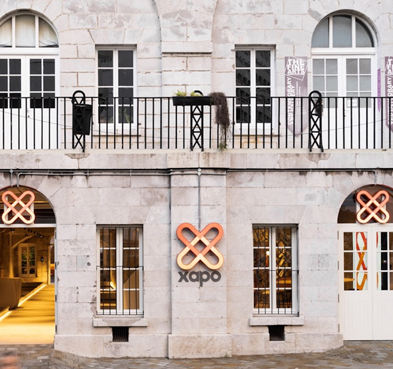 The exterior of the Xapo Bank building featuring a historic light-colored stone facade, arched doorways, grilled windows, and the orange and yellow infinity-loop Xapo logo mounted on the central pillar and above the entrances.
