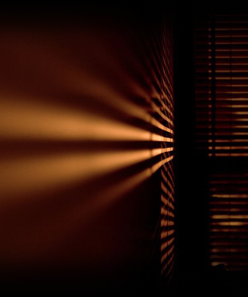 Low-key cinematic photography capturing dramatic chiaroscuro lighting, with warm orange light beams breaking through window venetian blinds into a pitch-black room.