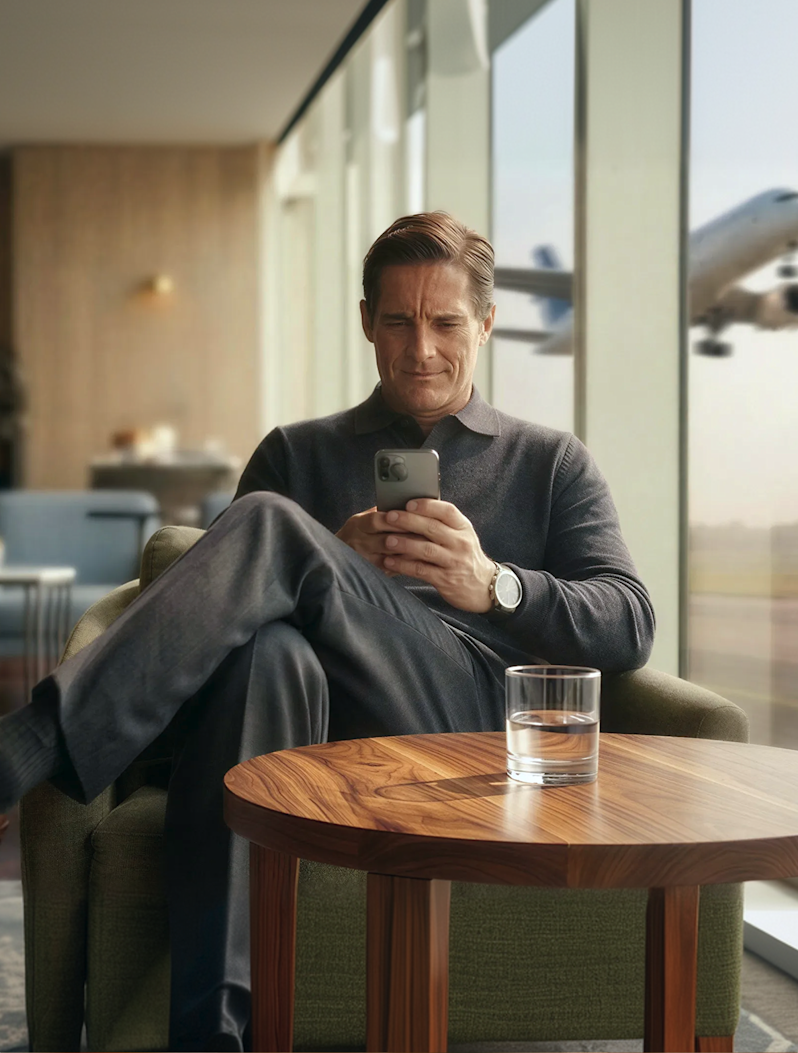 A man sitting in a lounge chair using his phone with an airplane taking off through the window behind him.