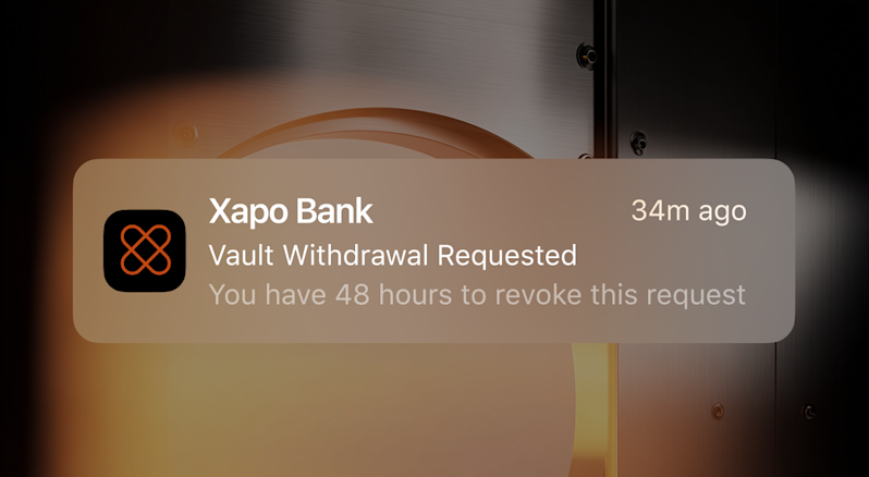 A close-up of a mobile push notification from Xapo Bank stating 'Vault Withdrawal Requested' and 'You have 48 hours to revoke this request', overlaying a background image of a metallic security vault door illuminated by a warm orange glow.